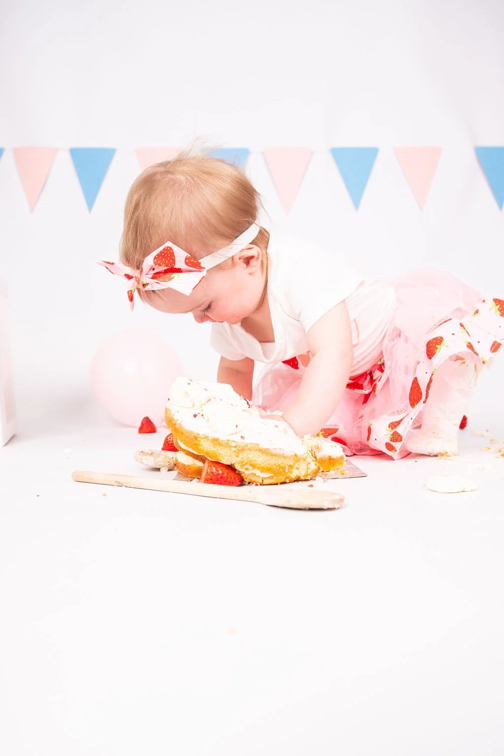 A young girl with blonde hair wearing a strawberry-patterned headband and pink tutu is playing with a cake with strawberries on it at a celebration with pink balloons and bunting.