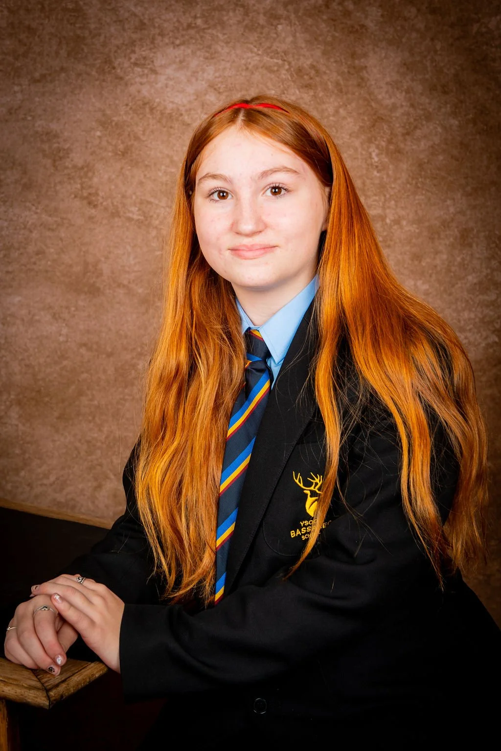 Portrait of a young woman with long red hair, wearing a school blazer with a crest, shirt, and striped tie, sitting at a table with hands resting on it, against a plain brown background.