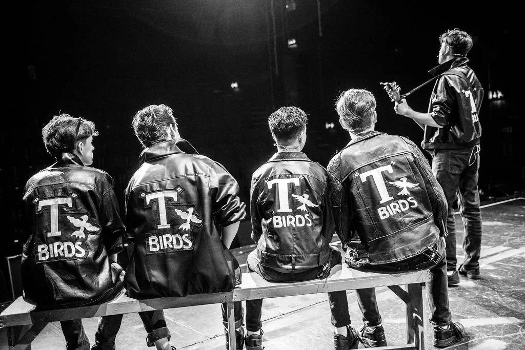 Five young men with leather jackets labeled 'T Birds' sitting on a bench, two of them are facing away, one is facing sideways, and two are turned slightly towards each other. They are watching one of their friends playing a guitar on stage in a dark 