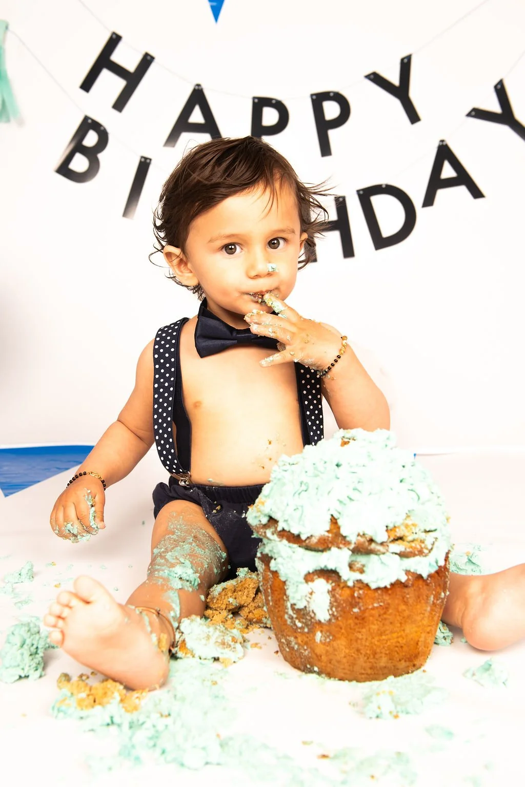 A young boy celebrating his birthday with a messy cake, wearing a black bow tie and suspenders, in front of a 'Happy Birthday' banner.