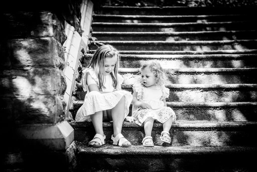Candid black and white photo of two little girls sitting on rustic stone steps in Belle Vue Park, Newport, captured by Emotion Picture Photography.