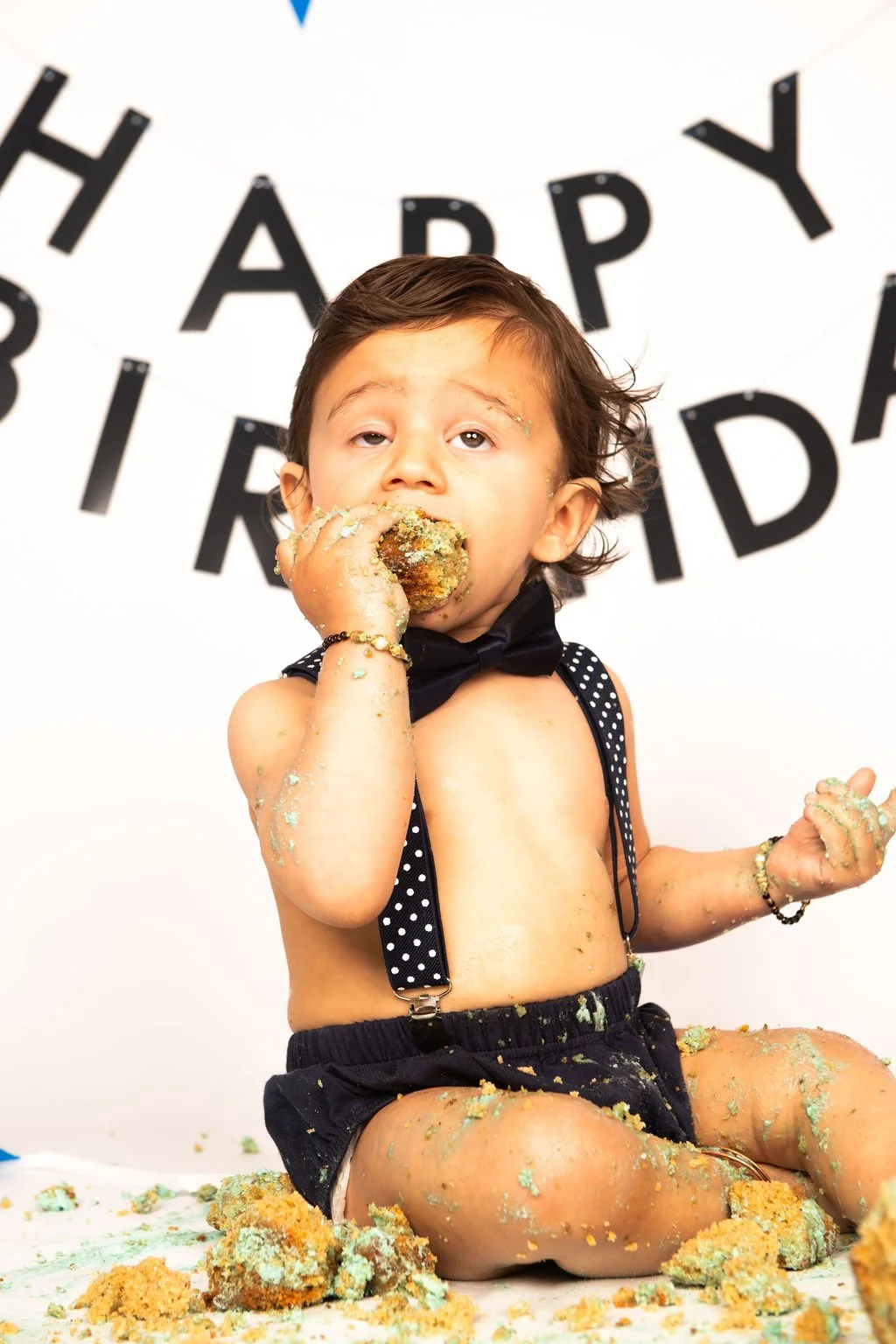 Young boy in black polka dot shorts and suspenders eating a cake, with cake and crumbs on his hands, face, and legs, in front of a 'Happy Birthday' banner.