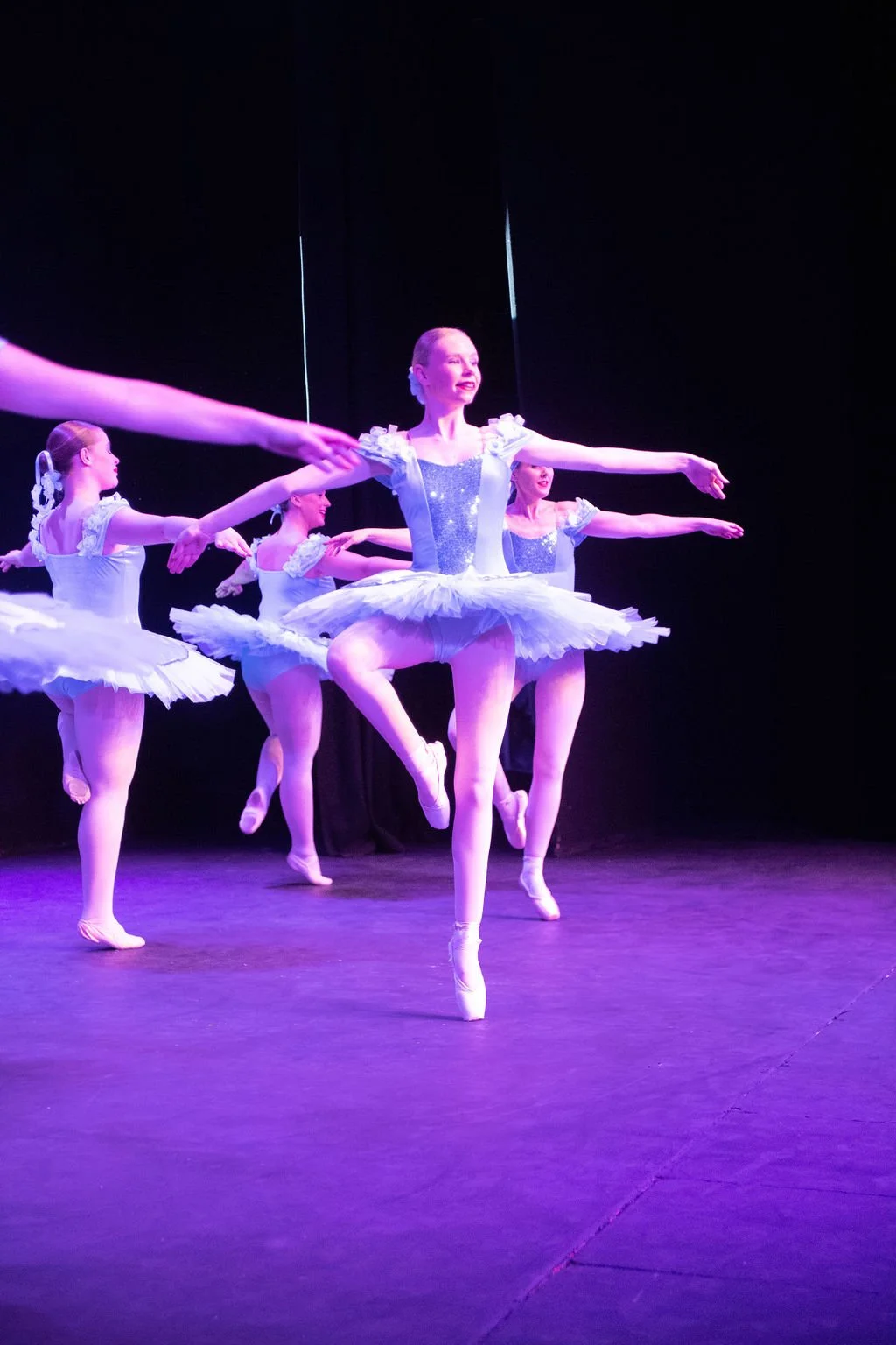 Ballet dancers performing on stage with purple lighting, dressed in white tutus and leotards.