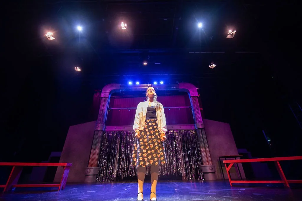 A woman standing on stage with colorful lighting and a curtain backdrop, possibly during a performance or rehearsal.