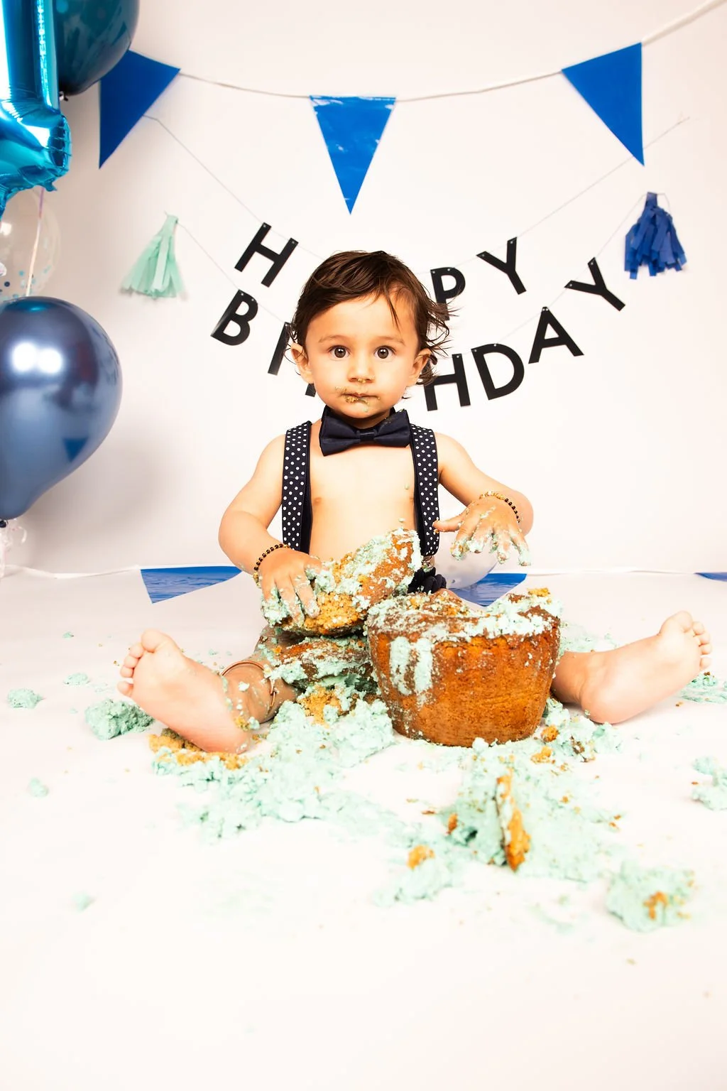 A young child with messy green birthday cake on their hands and face, sitting on the floor in front of a cake and birthday decorations, celebrating their birthday.
