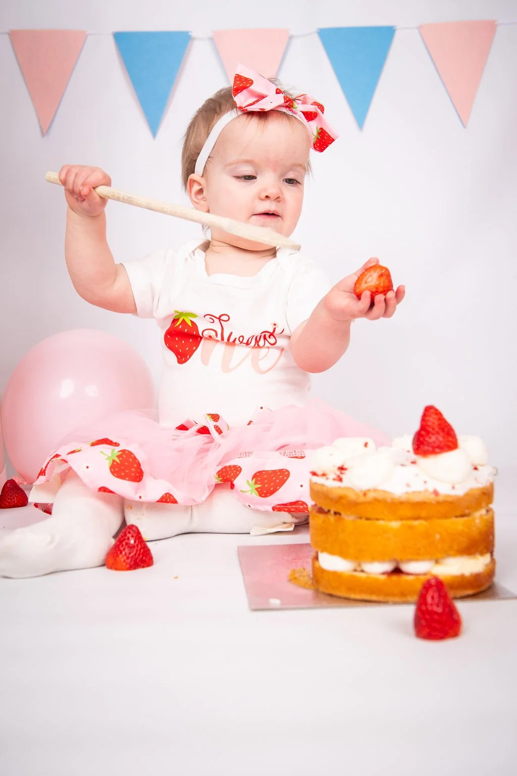 A young girl wearing a strawberry-themed outfit and pink bow headband, sitting on the floor with a pink balloon behind her. She is holding a strawberry in one hand and a wooden spatula in the other, in front of a strawberry shortcake decorated with s