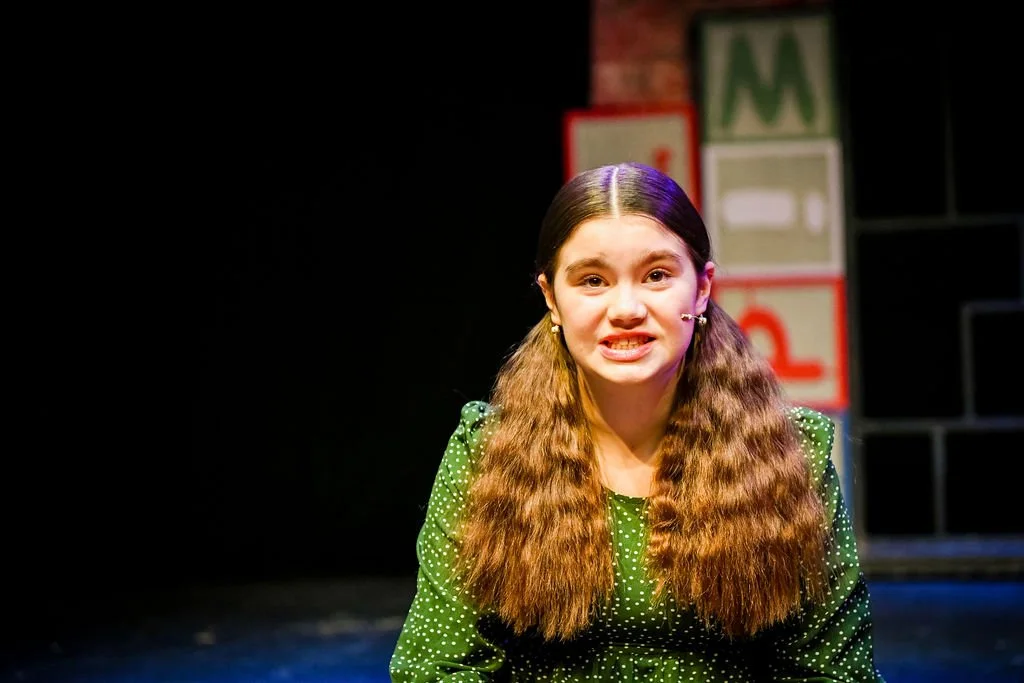 A young woman with long, wavy brown hair and braces, wearing a green polka dot dress, performing on stage with a background of colorful building blocks.