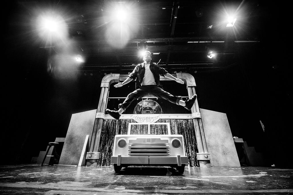 A performer jumps mid-air in front of a stage set resembling a vintage car, with a backdrop and stage props in a theater or concert setting.