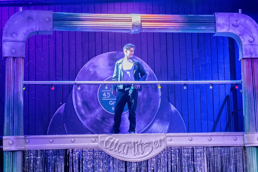 A person standing on a stage with a large, decorative circular backdrop behind them that resembles a vinyl record. The stage is framed with metallic and wooden elements, and colorful string lights are hanging across the front.