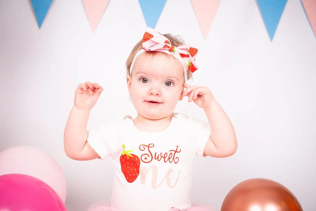 A young girl with a strawberry-themed headband and a white shirt with a strawberry and the words 'Sweet One' stands in front of colorful decorations, including pink, brown, and blue balloons and triangular banners.