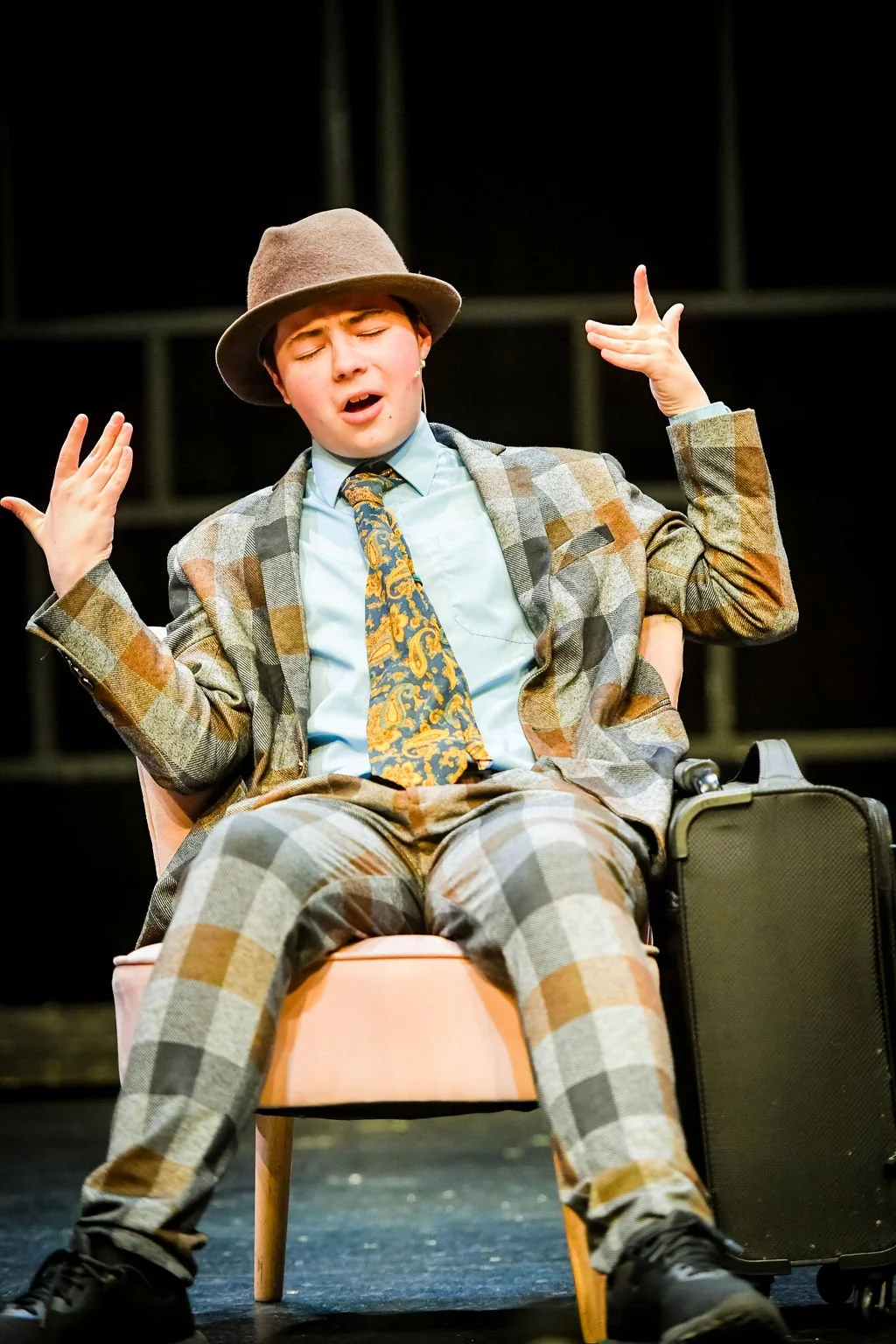 A young person in vintage-style clothing and a hat, appearing to perform or act on stage, sitting in a pink chair with a suitcase beside them.