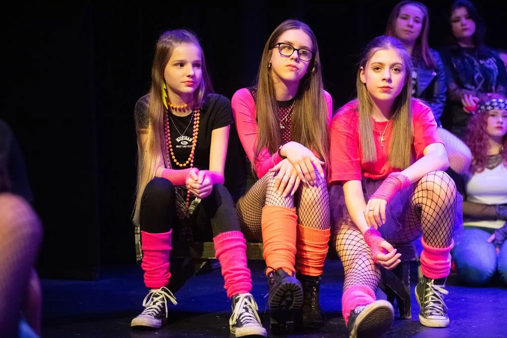 Three young girls sitting on a bench on stage during a performance, dressed in colorful 80s-inspired outfits with fishnet stockings and leg warmers.