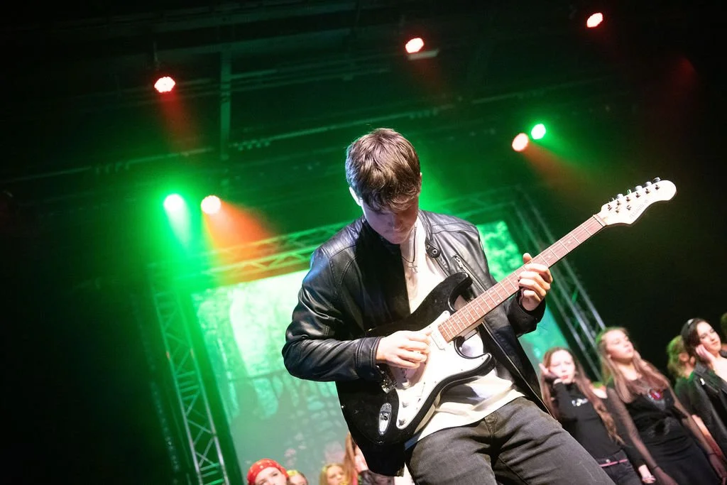 A young man playing an electric guitar on stage, with several women standing behind him, colorful stage lights, and a large screen in the background.