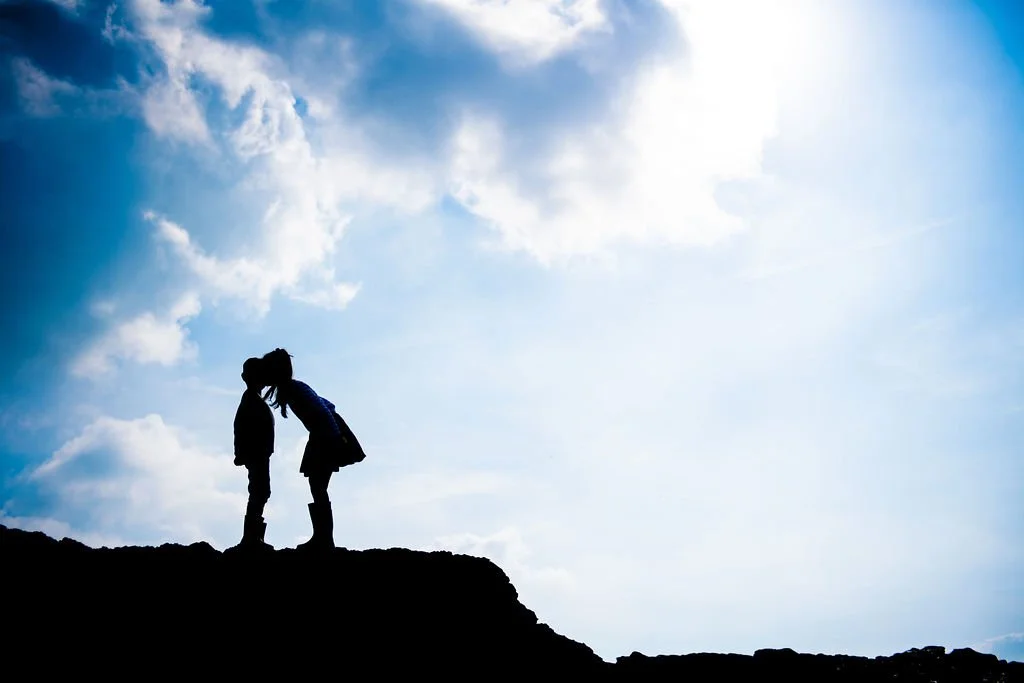 Silhouette of two children, a boy and a girl, standing on a rocky ledge and touching foreheads against a bright sky with clouds.