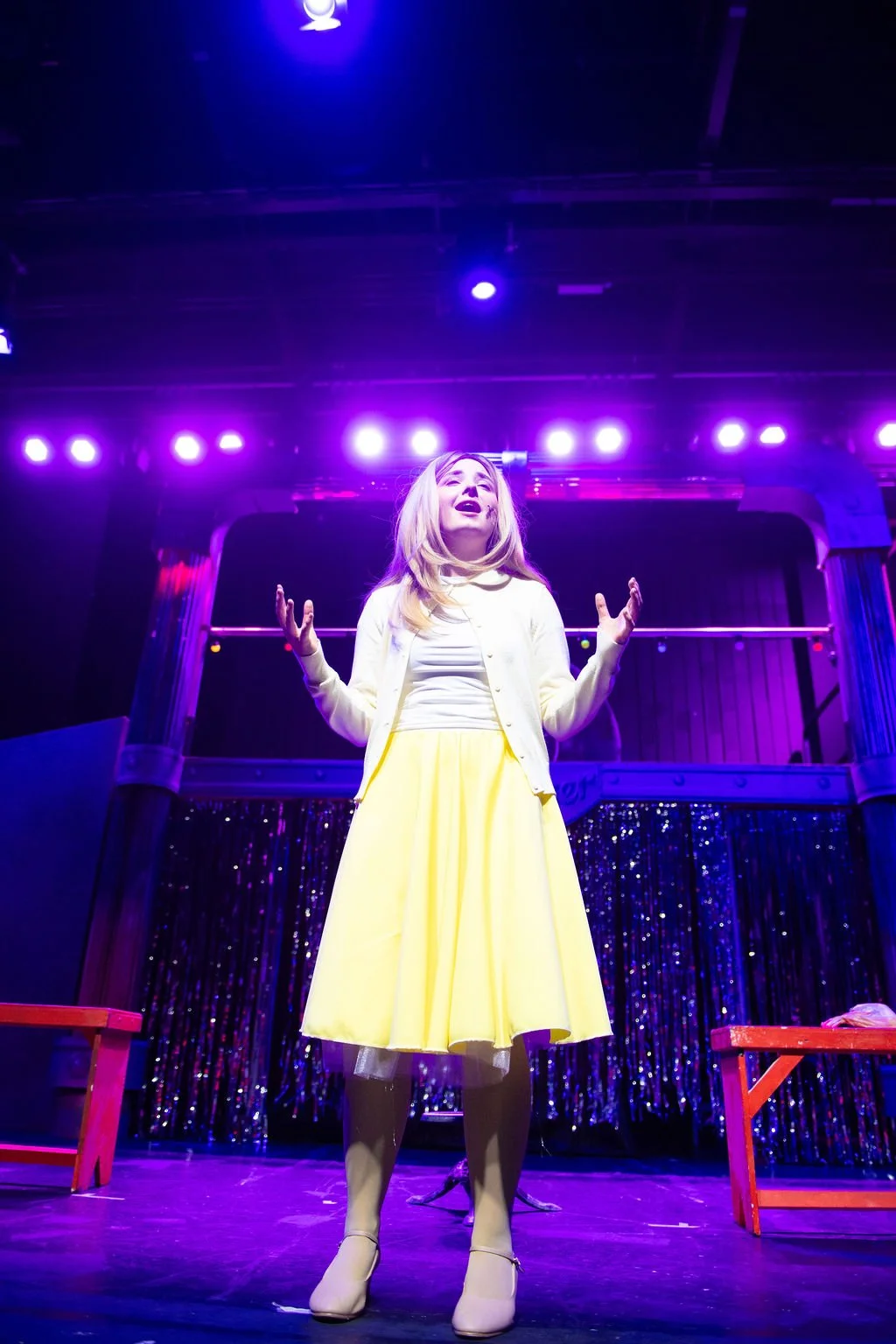 Young woman on stage during a theatrical performance, under purple stage lights, wearing a yellow skirt, white top, and beige boots.