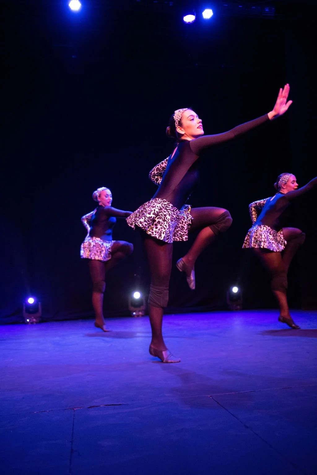 Three dancers performing on stage with black background and purple lighting, dressed in black dance leotards with patterned skirts and headbands.