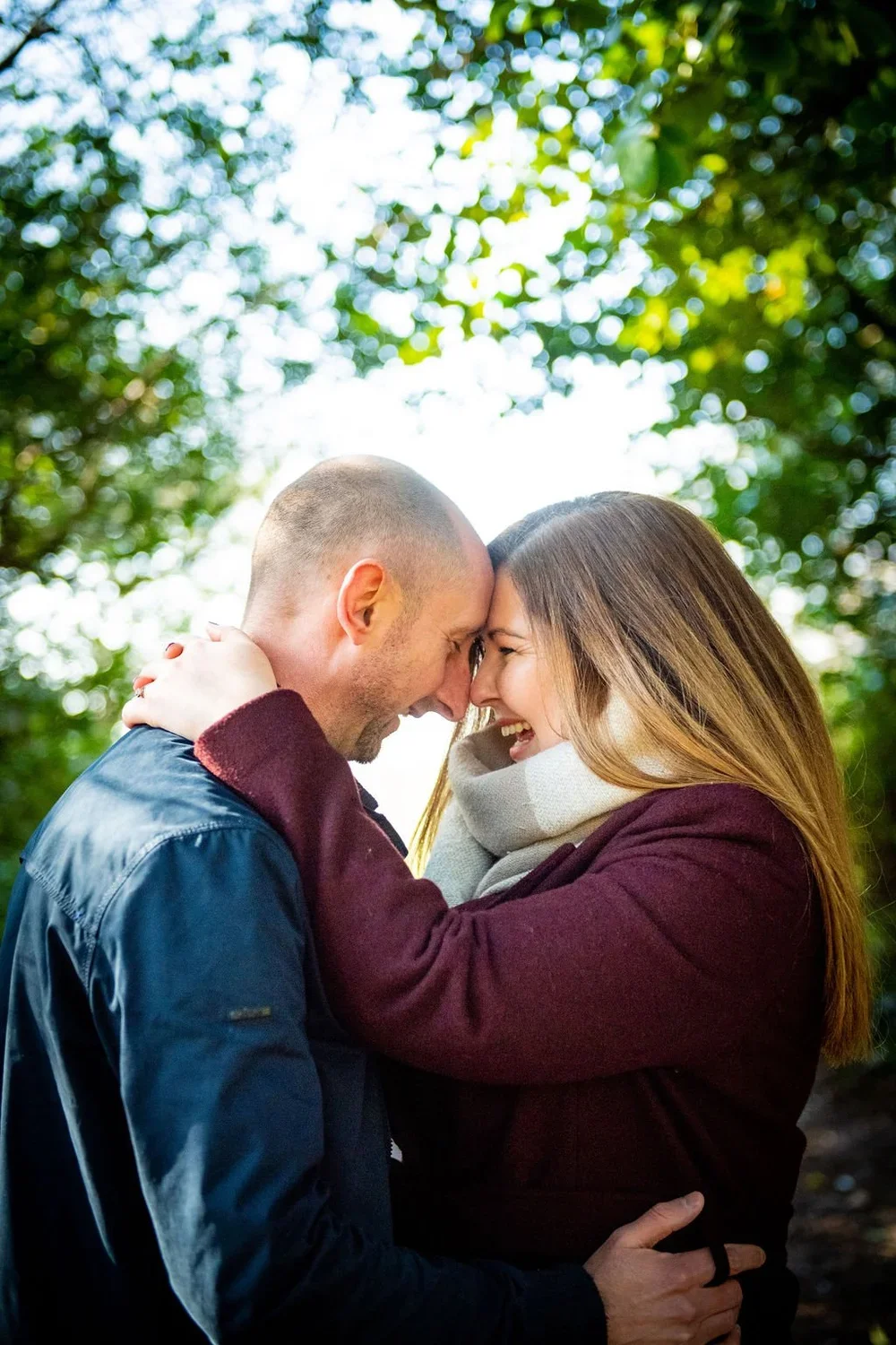 A couple with their foreheads and noses touching, smiling joyfully in an outdoor setting with green trees and bright sky behind them.