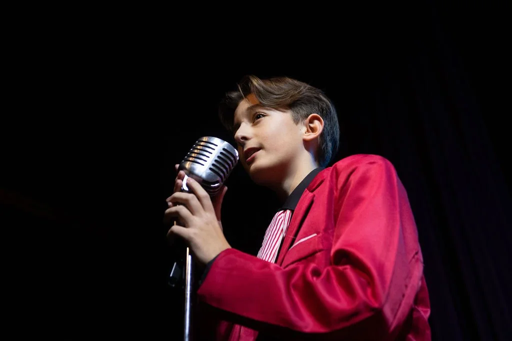 A young boy in a red jacket speaking into a vintage microphone on a stage with a black background.