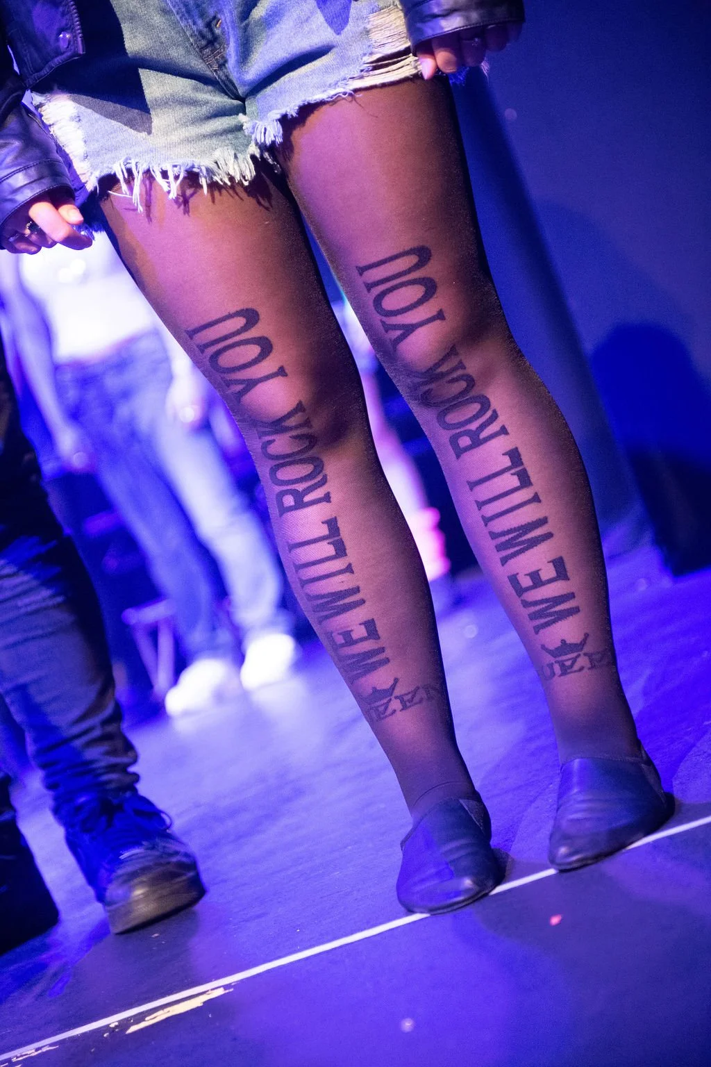 Legs of a person with black tights that have the phrase "EXILE WILL ROCK YOU" written on them in large black letters. The person is wearing denim shorts and black shoes, standing on a dark stage with purple lighting.