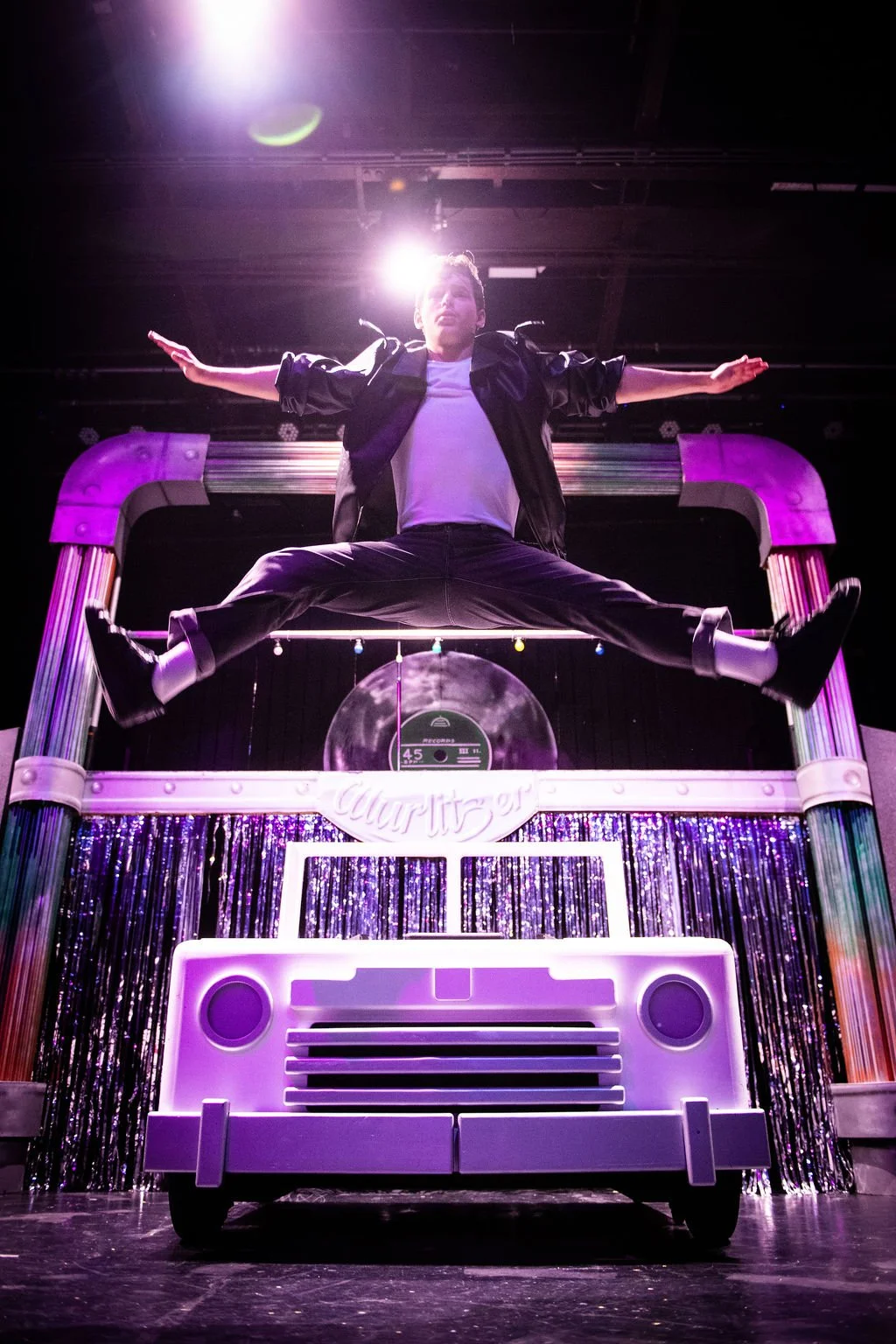 A man is performing a jump in mid-air on a stage decorated with colorful neon lights and a large boombox prop, with purple and pink lighting effects.