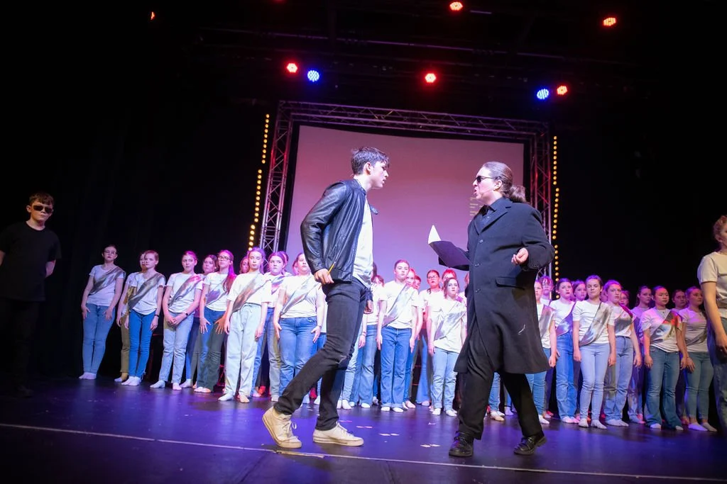 A stage performance with a youth choir, a man and a woman in conversation, stage lighting, and a large screen in the background.