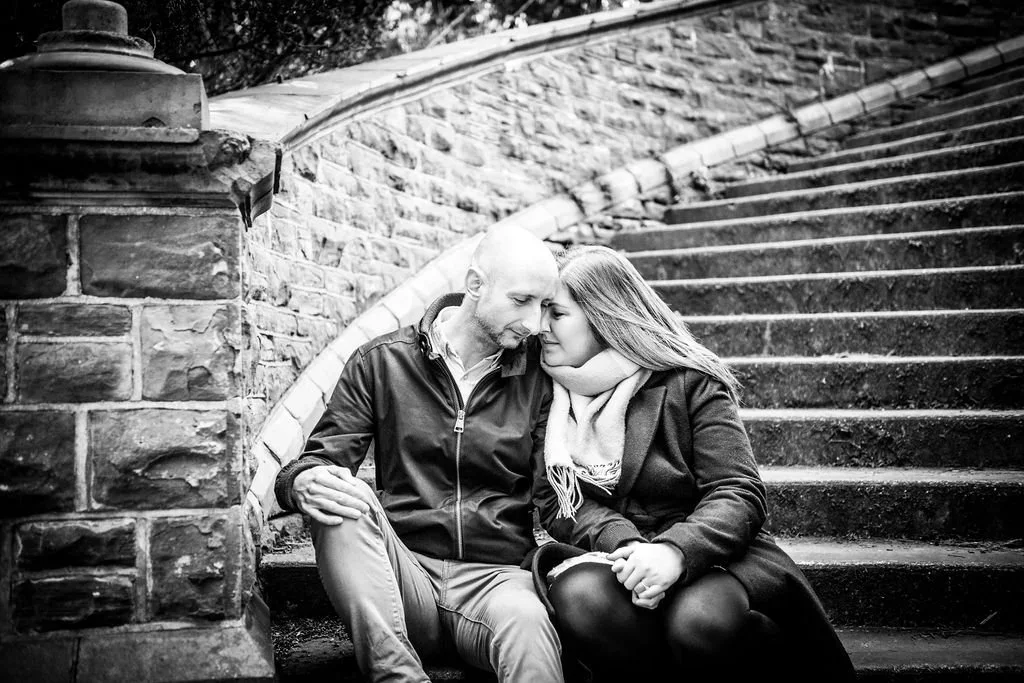 A black and white photo of a couple sitting on outdoor stairs, leaning their foreheads together in an intimate moment.