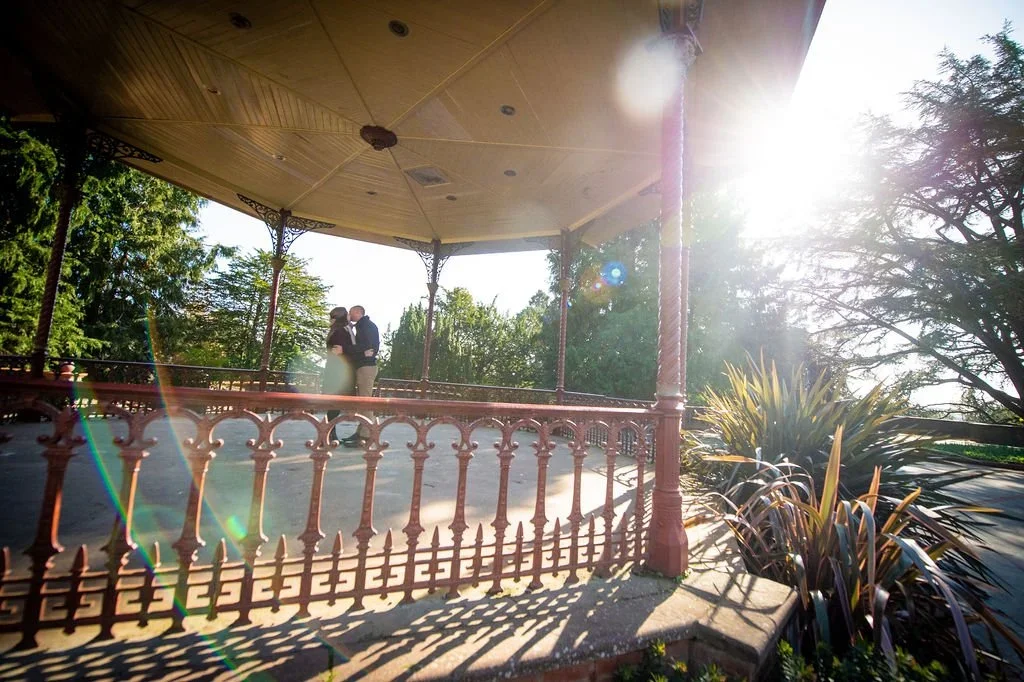 A couple dressed in formal attire sharing a kiss on a gazebo in a park on a sunny day with trees and plants around.
