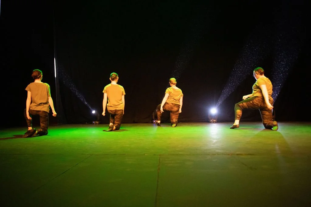Four people kneeling on a stage with green lighting, facing black curtains, with stage lights shining behind them.