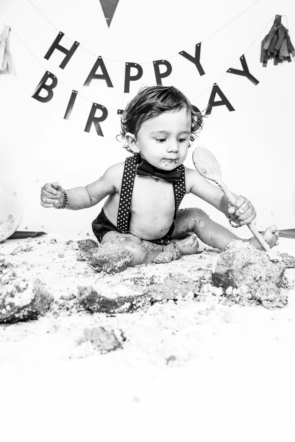 A child with curly hair, wearing suspenders and a bow tie, sitting on the floor covered in sand, playing with a sand shovel. There is a 'Happy Birthday' banner in the background, indicating it is a birthday celebration.