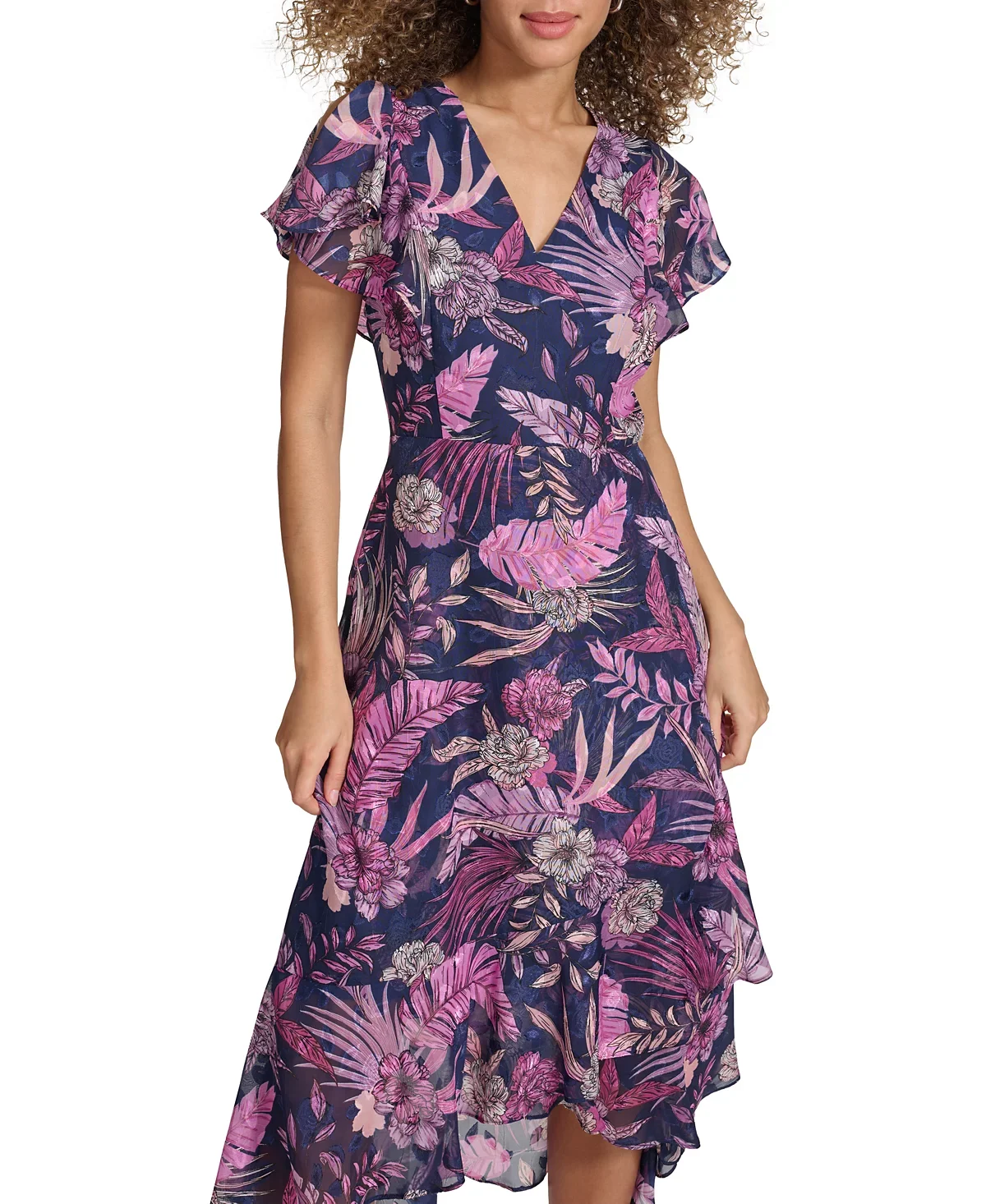 A woman wearing a long, flowy dress with a floral and leaf print in shades of pink, purple, and beige on a dark blue background, standing against a plain white background.