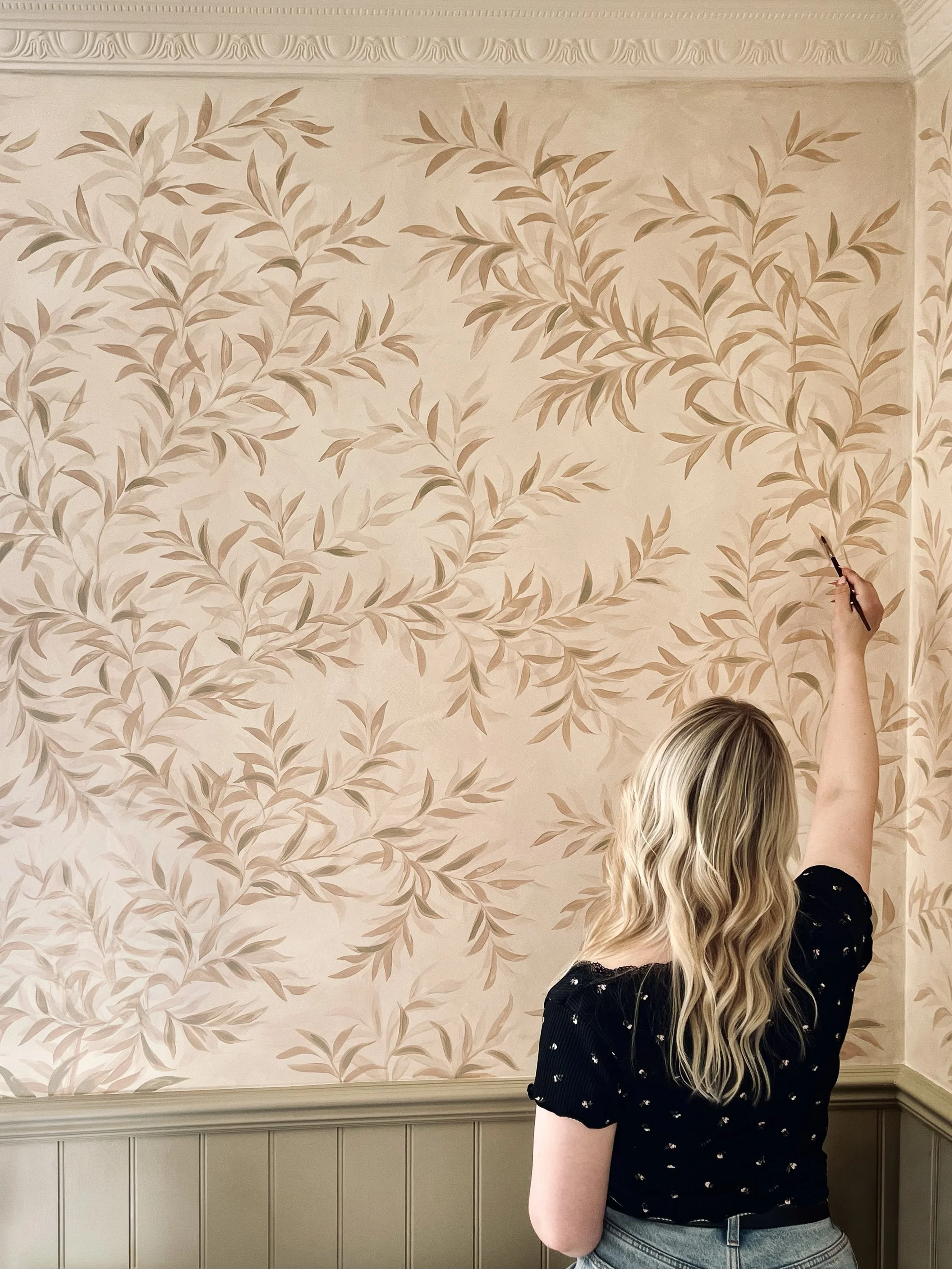 A woman with wavy blonde hair is painting a floral pattern on a wall with beige and pink leaves, wearing a black dress with small white flowers and jeans.