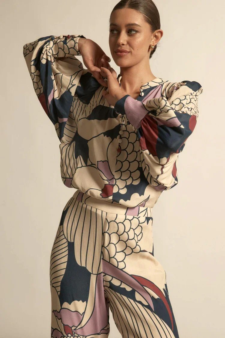 A woman with brown hair tied back, wearing a matching floral print blouse and pants with a beige background and large dark navy, pink, and red floral patterns. She is posing with her left arm bent and hand near her face against a plain beige backgrou