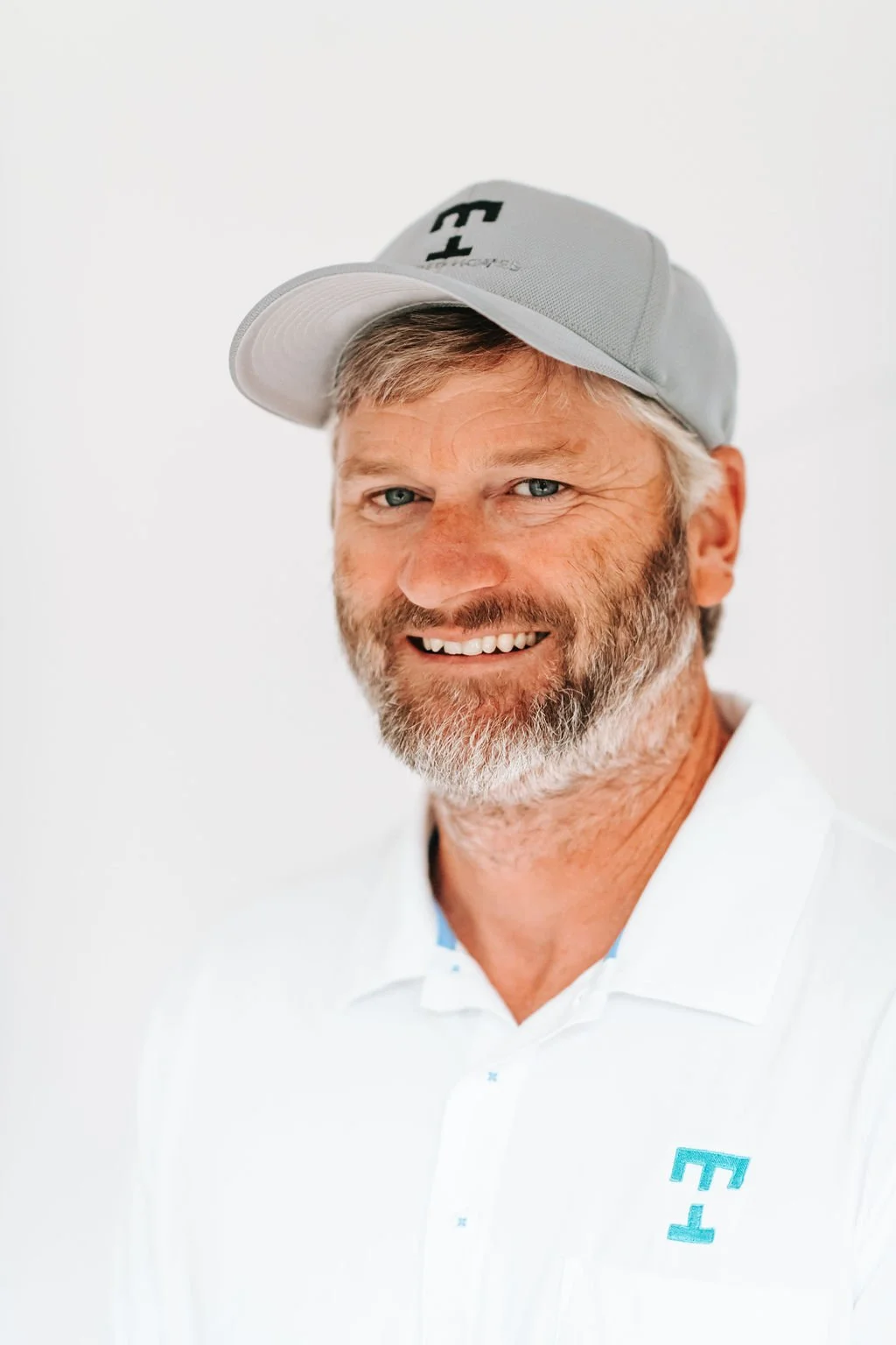 Smiling man with gray hair and beard standing at the doorway, wearing a navy blue polo shirt with Under Armour logo and a cap.