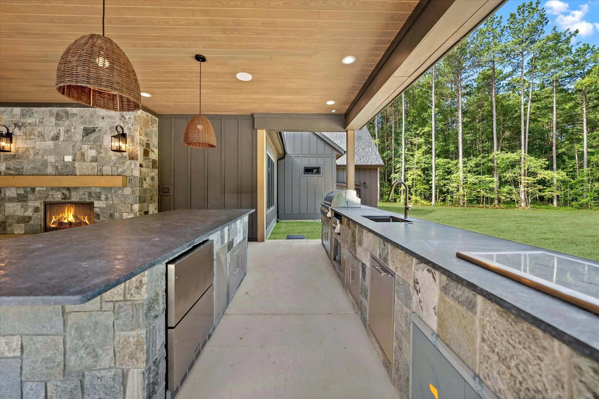 Outdoor kitchen with stone countertops, a built-in grill, a sink, and a view of a wooded backyard.