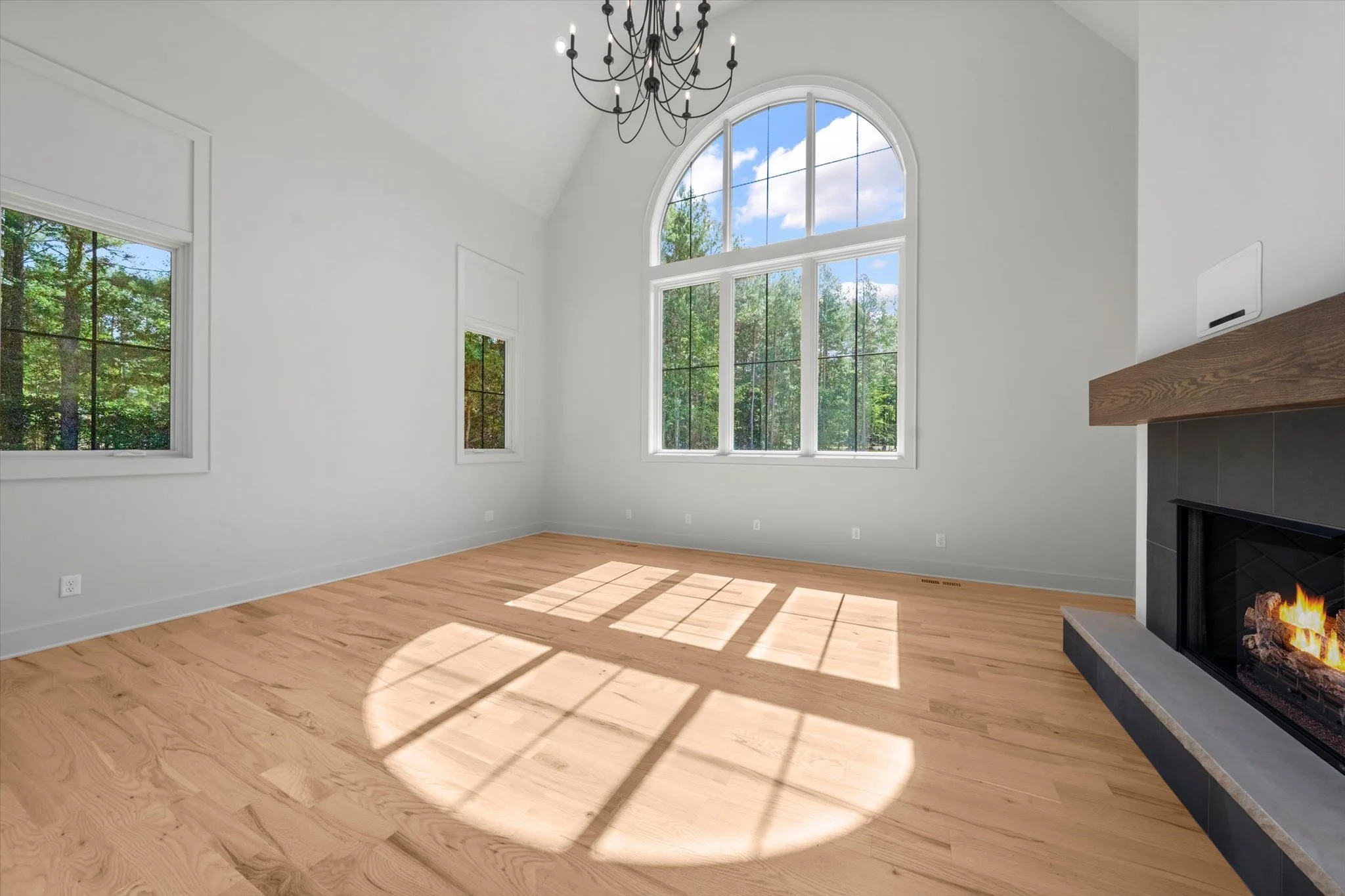 Empty living room with large arched windows, hardwood floors, a black chandelier, and a fireplace with a wood mantle and fire burning inside.