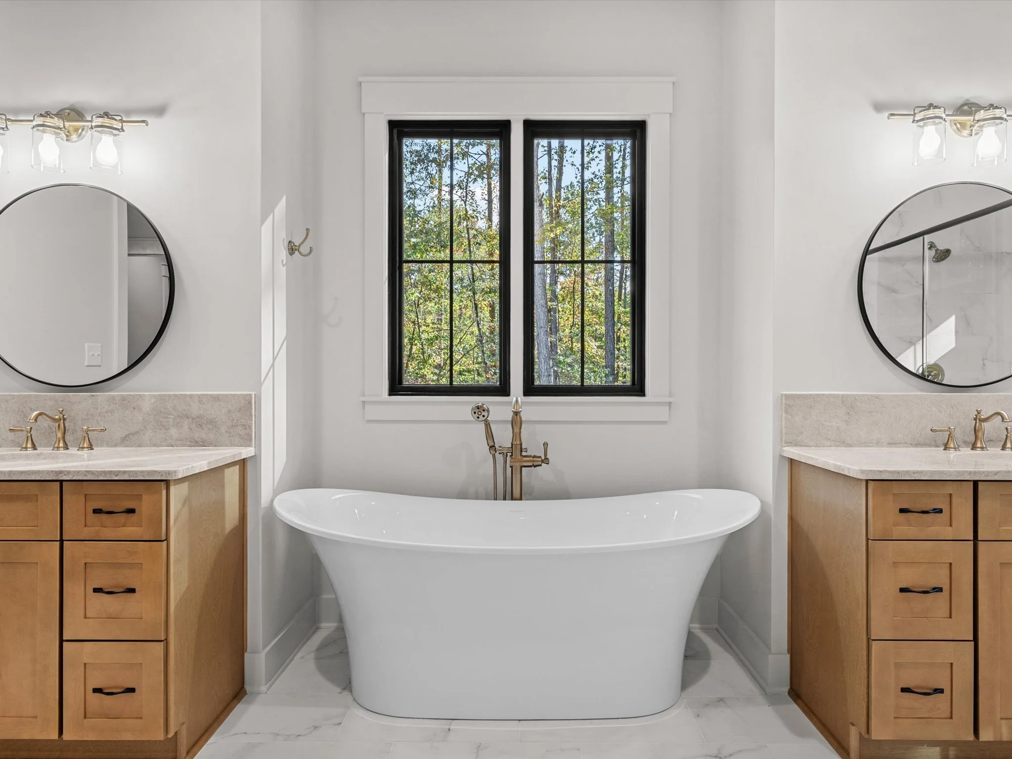 A modern bathroom featuring a white freestanding bathtub centered beneath a black double-pane window with a view of trees outside. The bathroom has two matching wooden vanities with marble countertops, each with an oval mirror and brass fixtures, fla