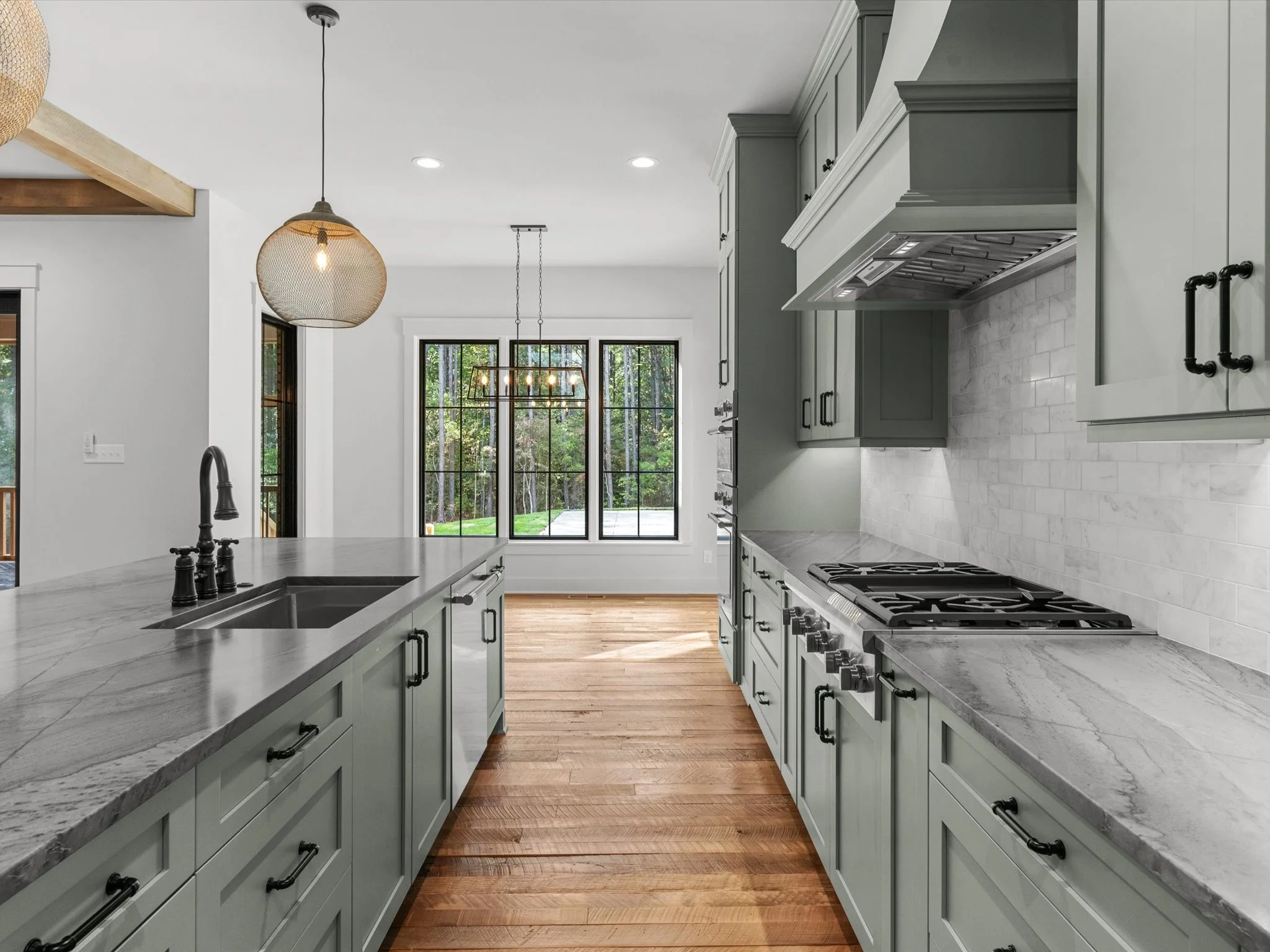 Modern kitchen with light grey cabinets, marble countertops, black hardware, gas stove, white brick backsplash, hardwood floors, and large windows overlooking a green outdoor area.