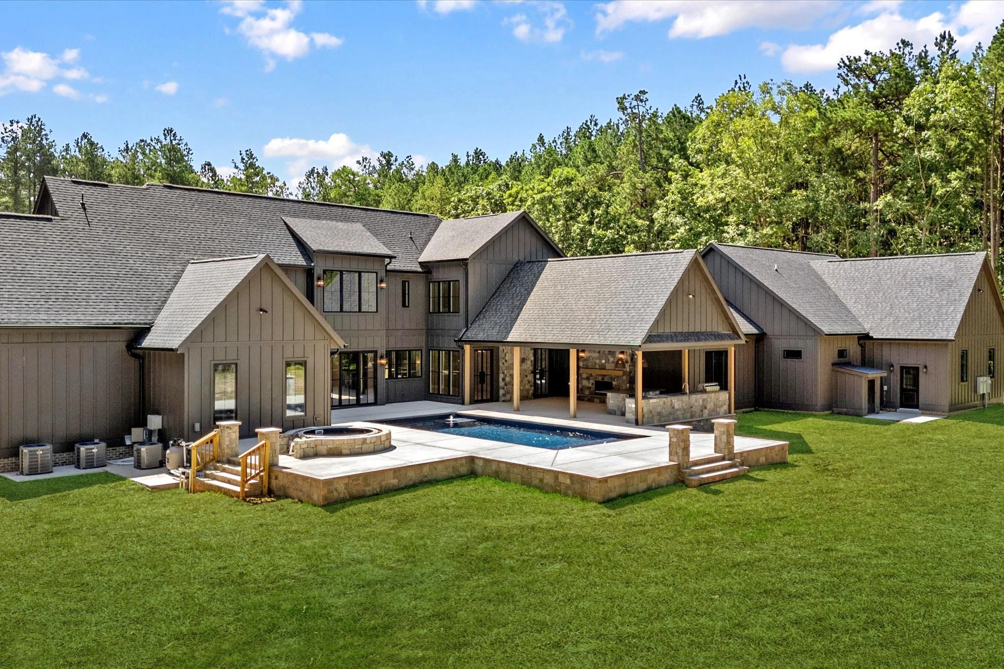 A modern house with a backyard featuring a pool, hot tub, and outdoor kitchen, surrounded by green lawn and trees.