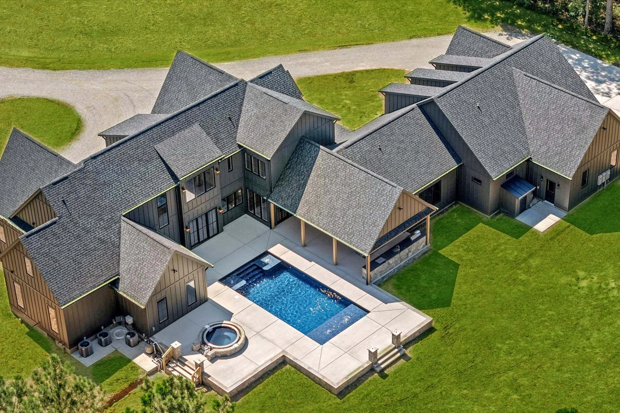 A large modern house with multiple gabled roofs, gray shingles, and dark siding, featuring a backyard with a rectangular swimming pool, a hot tub, and a spacious patio surrounded by green lawn.
