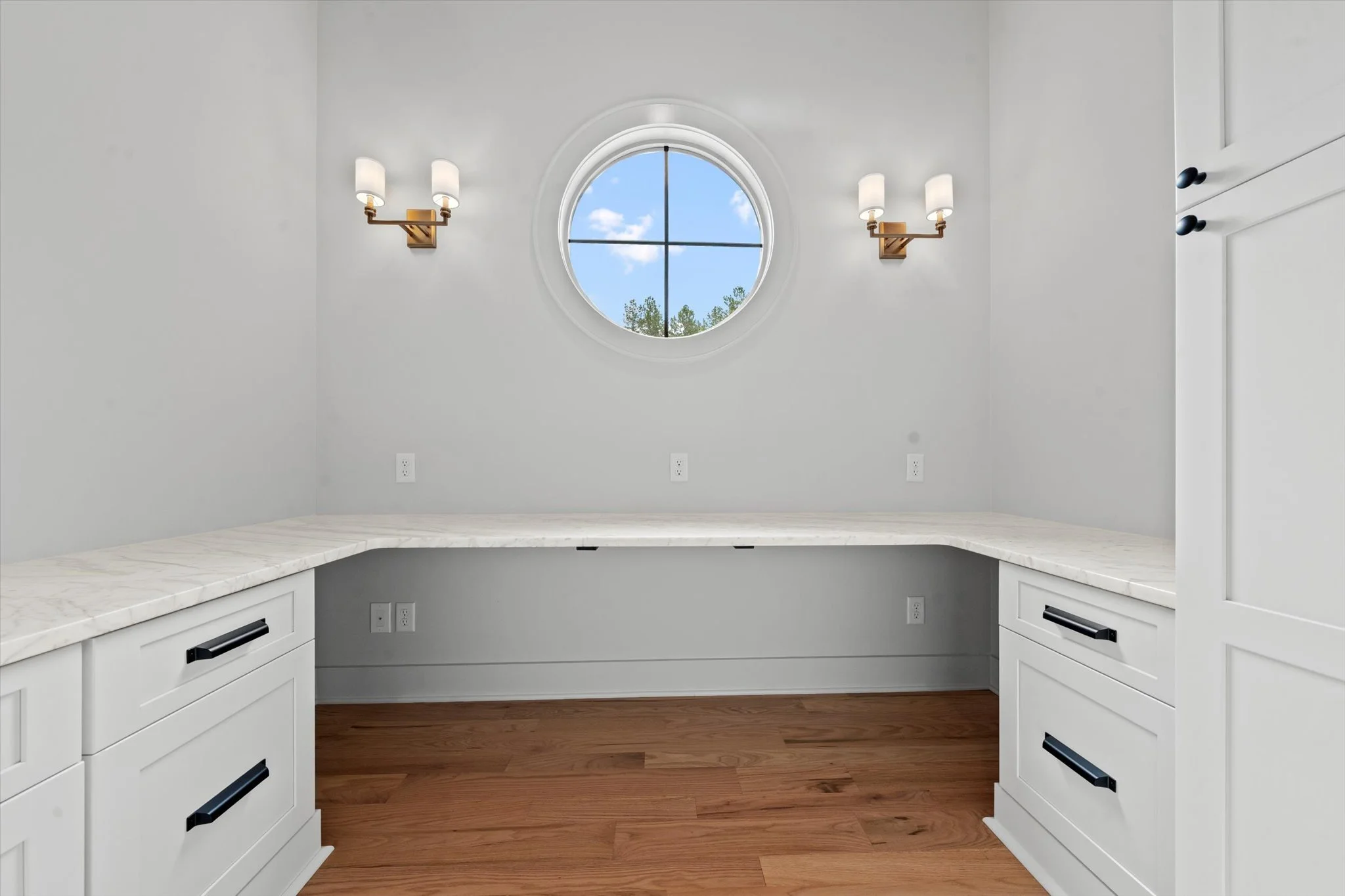 Empty kitchen nook with white cabinetry, marble countertop, hardwood floor, two wall sconces, and a circular window showing blue sky and clouds.