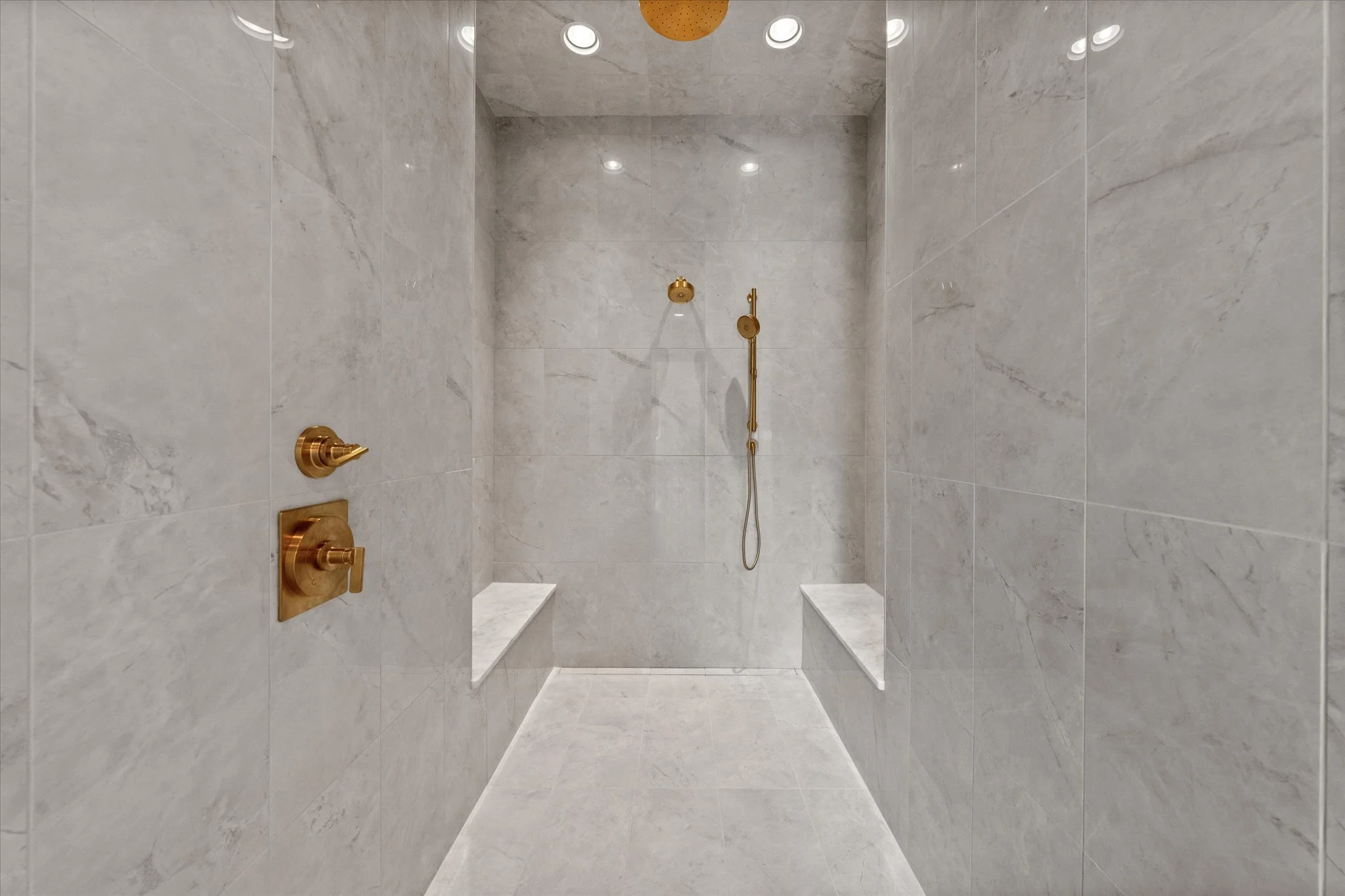Empty walk-in shower with beige marble tiles, two built-in benches, gold fixtures including a showerhead, handheld shower, and water controls, and three ceiling lights.