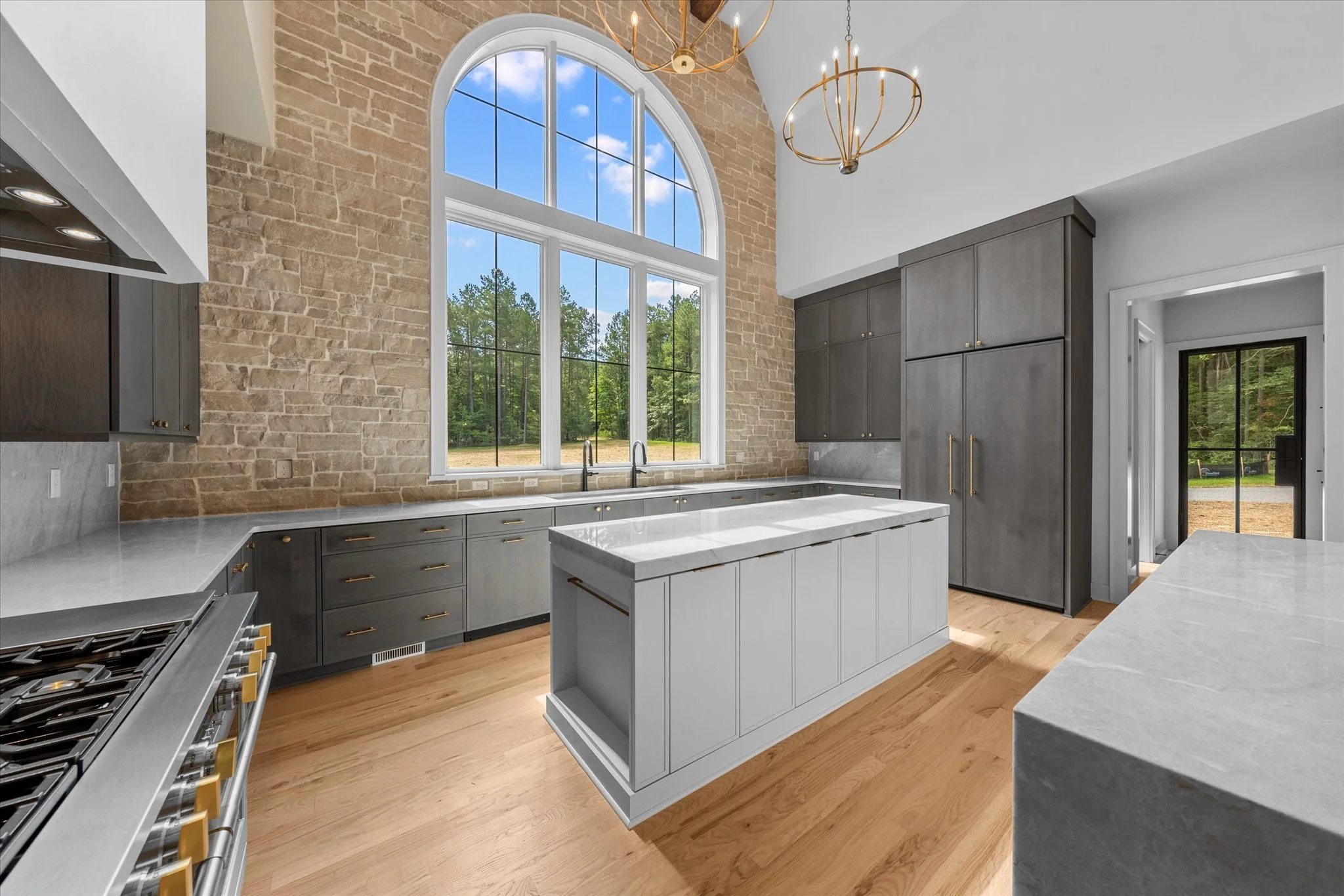 Modern kitchen with large arched window, light wood flooring, gray cabinets, marble countertops, and a brick accent wall, with two gold chandeliers hanging from the ceiling.