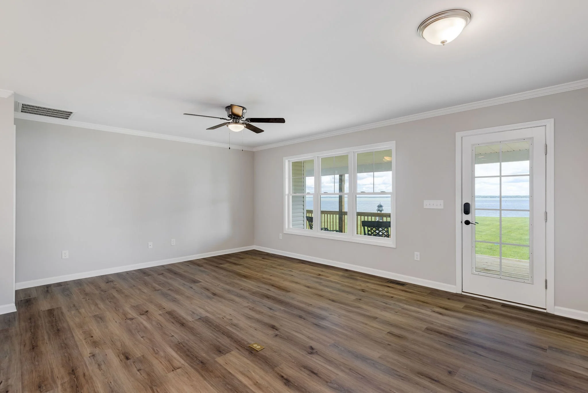 Empty living room with large window and glass door leading to a deck, overlooking a lawn and body of water, with wooden flooring, ceiling fan, and white walls.