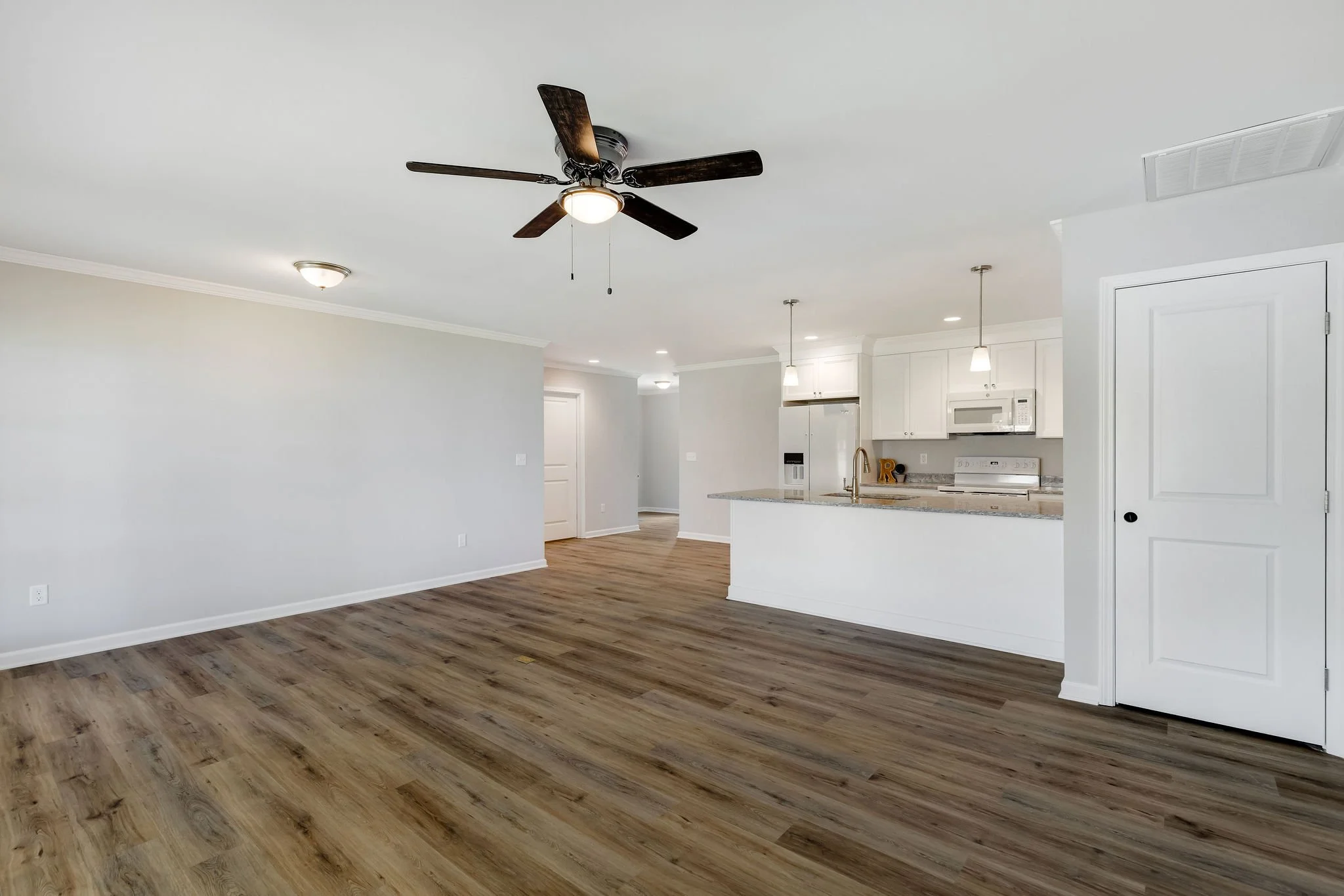 Empty open-concept living space with hardwood floors, white walls, ceiling fan, and a kitchen with white cabinets, granite countertops, and modern appliances.