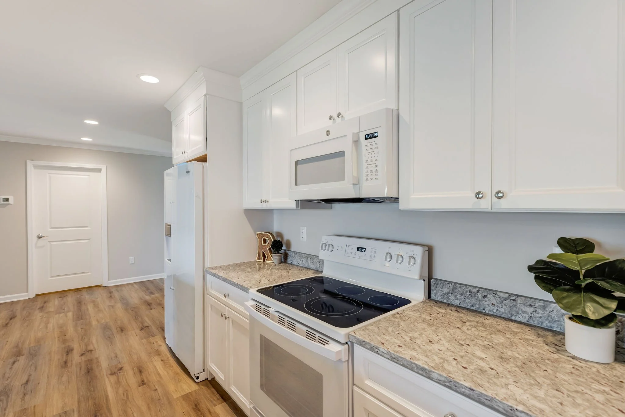 Kitchen with white cabinets, granite countertops, and white appliances including microwave and stove. Wooden flooring, decorative letter 'R' and potted plant on the counter.