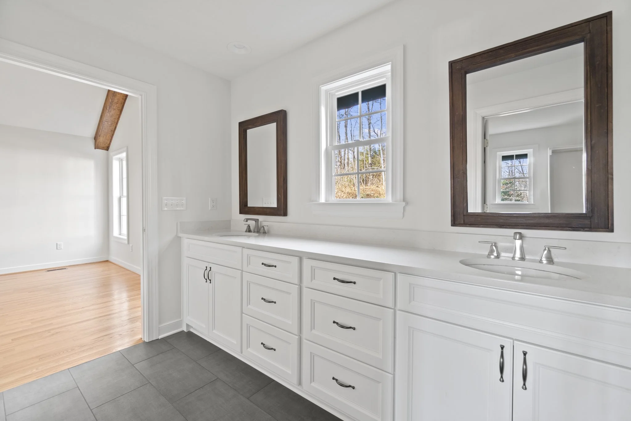 Bathroom with white cabinetry, two square mirrors with wooden frames, and two sinks, with a window showing an outdoor landscape.