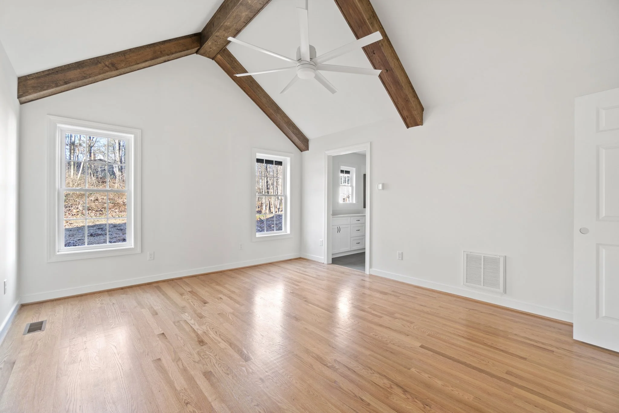 Empty room with light wooden floors, white walls, two windows, and a bedroom door. Exposed wooden beams and a white ceiling fan are on the ceiling.