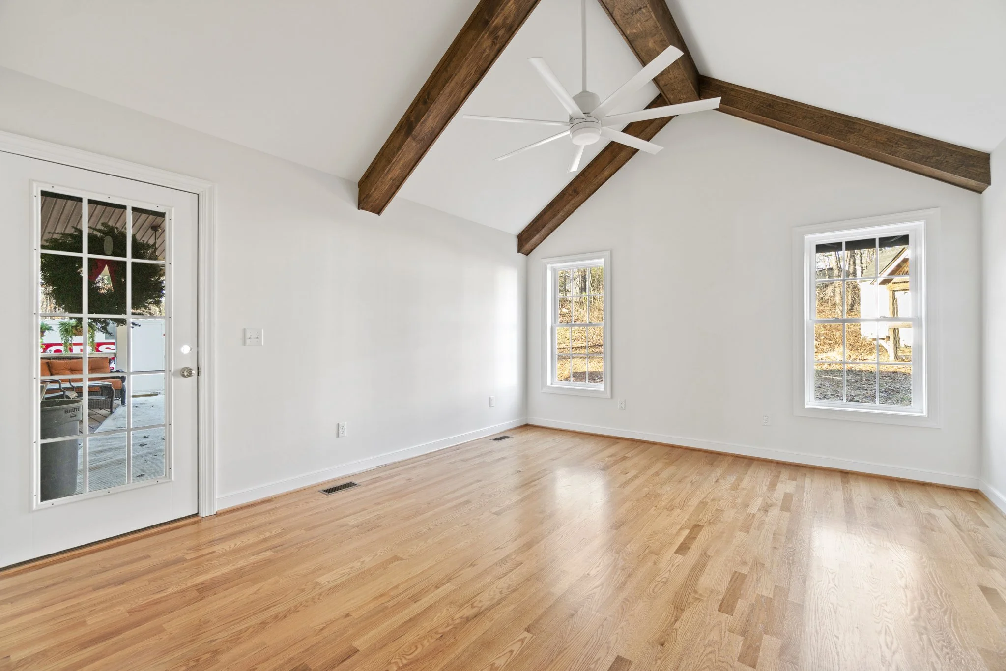 Empty room with white walls, wooden floors, two windows, a ceiling fan, and a door with glass panes leading outside.