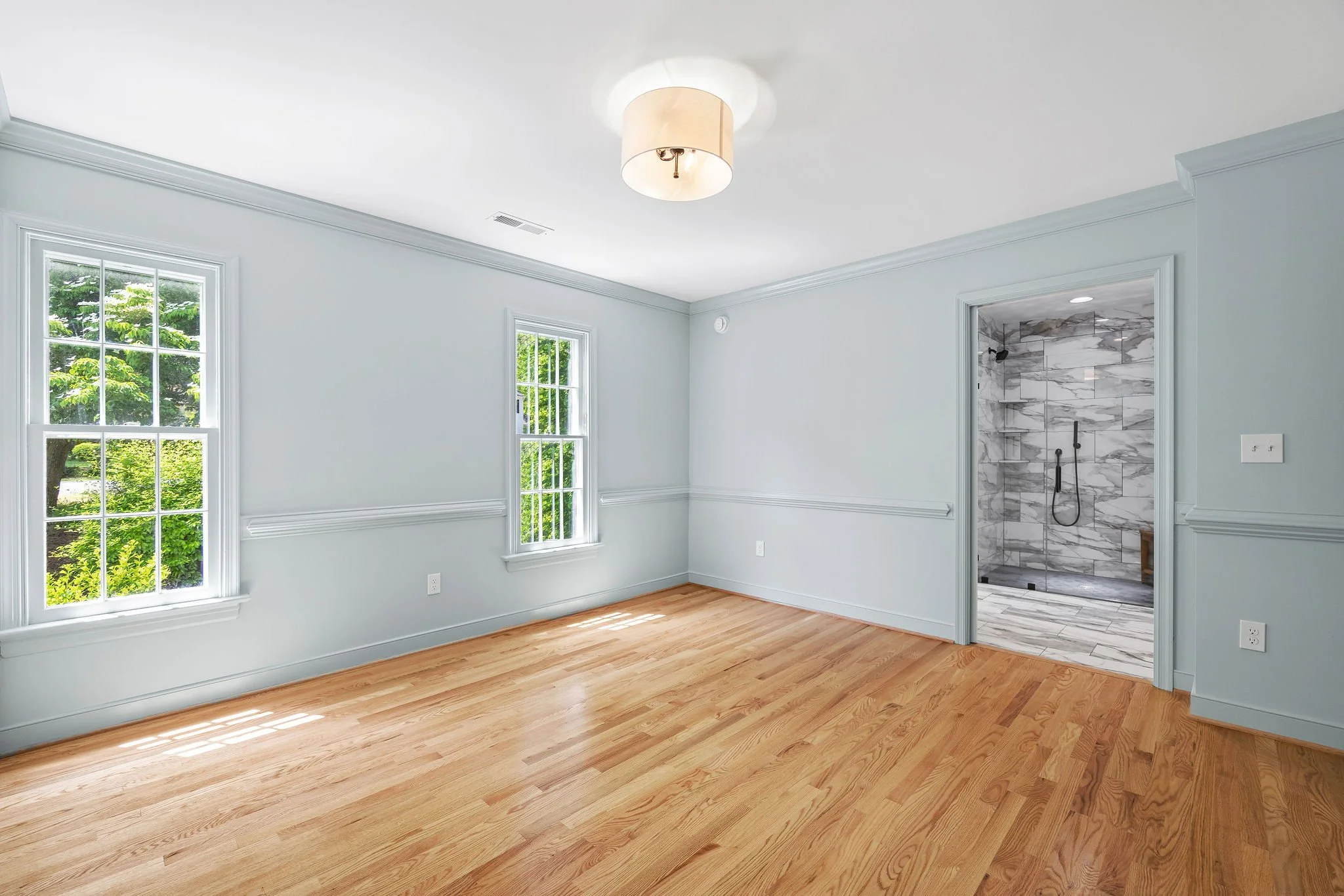 Empty room with two windows, hardwood floor, light-colored walls, and an open door to a marble-tiled bathroom or shower area.