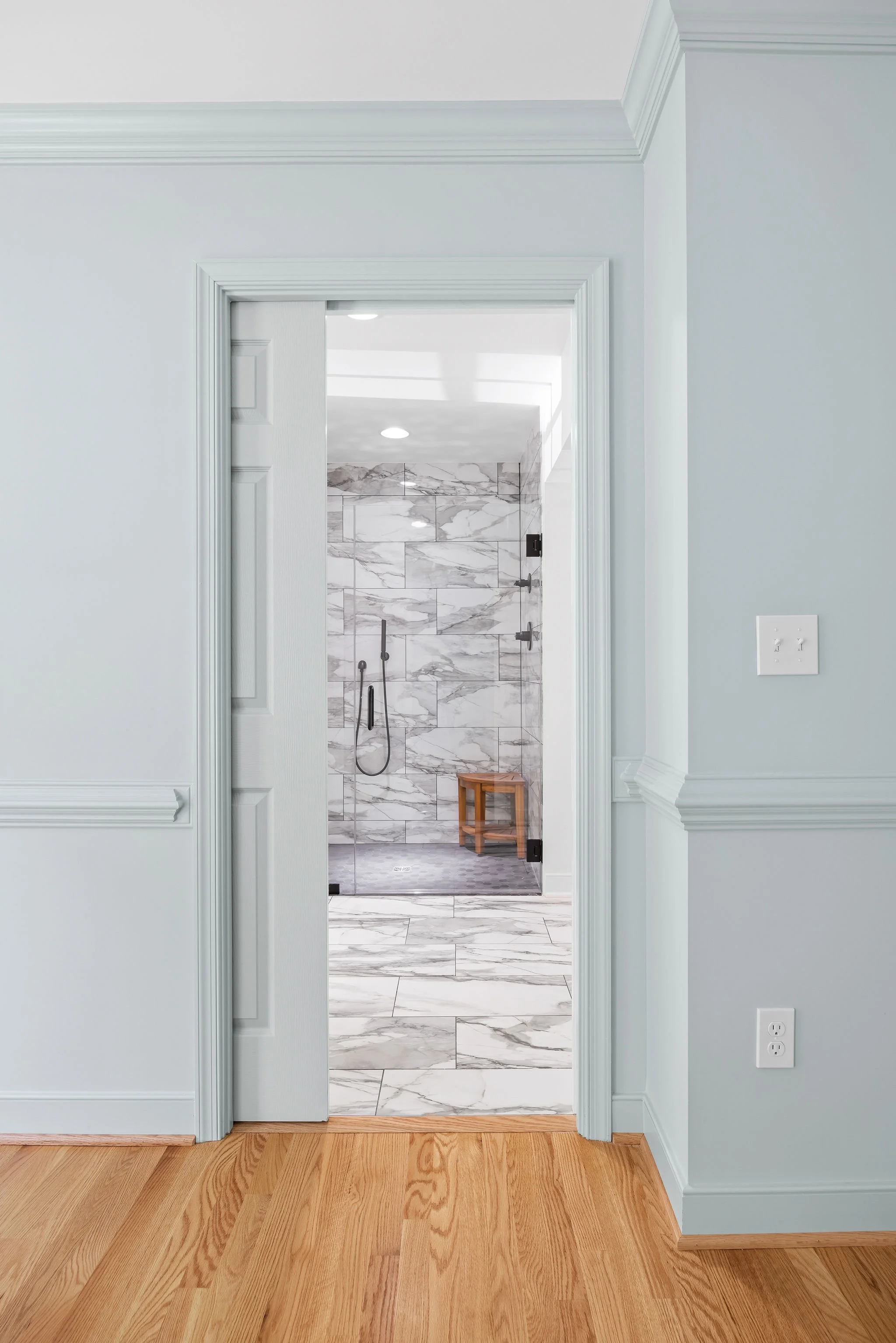 View of a modern bathroom through an open door, featuring a walk-in shower with marble tiles, a wooden stool, and gray and white color schemes.